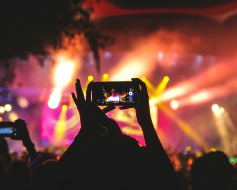 person holding smartphone taking video of a concert near stage with lights during nighttime