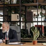 A senior man in a suit working at a desk in a modern office with plants.