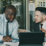Two businessmen collaborating over a tablet and laptop